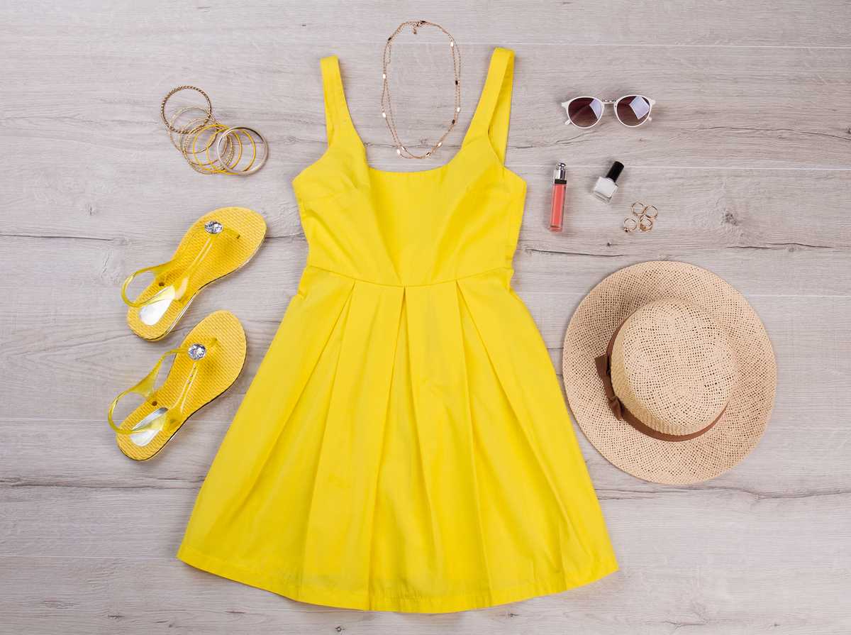 A yellow summer dress, yellow sandals, bracelets, a sun hat, some sunglasses and some makeup laid out on a wooden floor